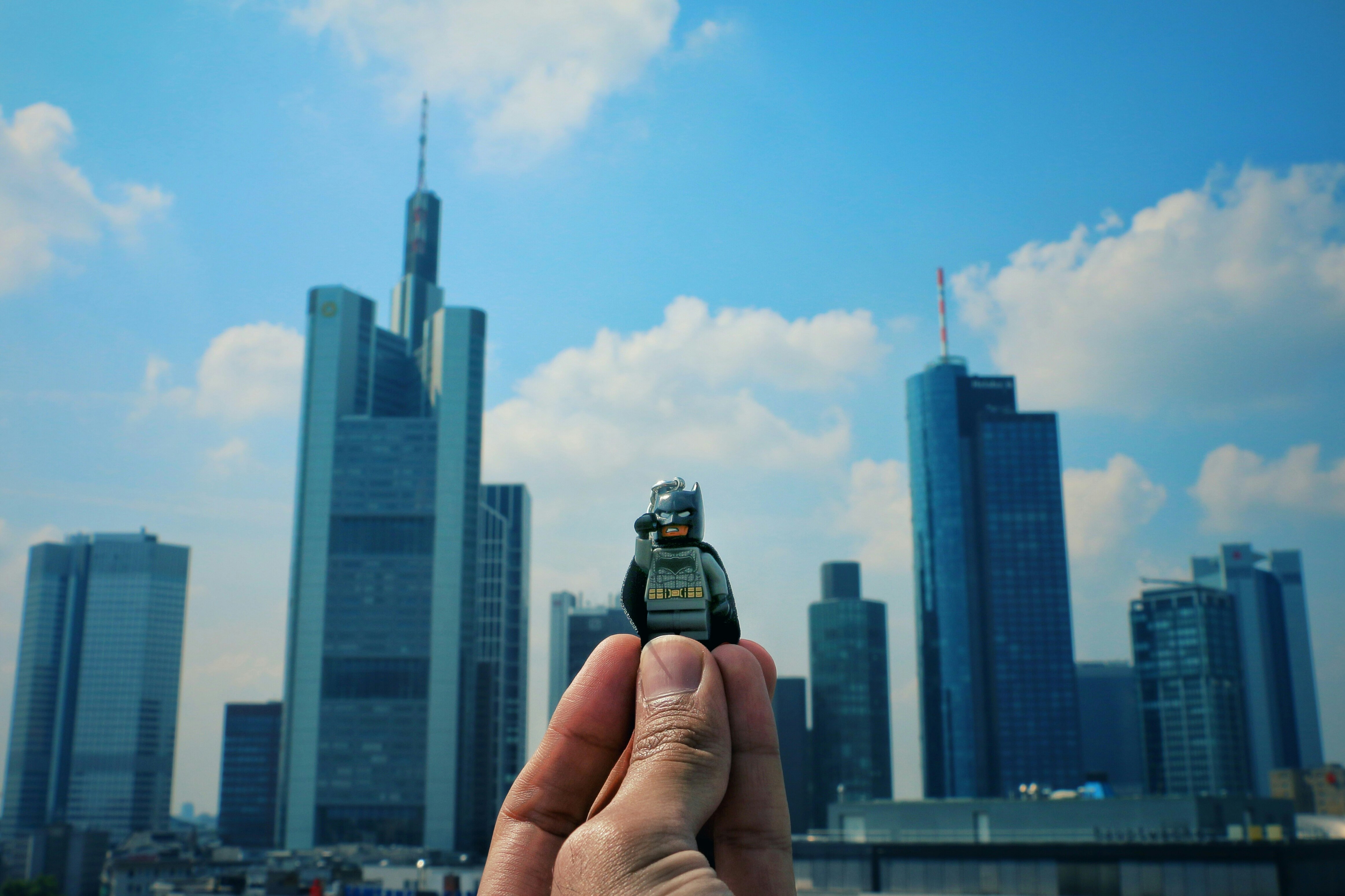 A person holding a Lego guy toy in the air with his fingers, with skyscrapers and a cloudy sky in the background.